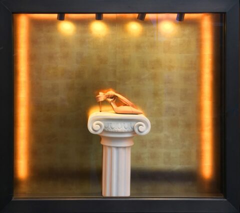 Schaufenster mir einer antiken Säule in der Mitte und einem Schuh darauf, die Rückwand ist mit einem gold gemusterten Dekor verkleidet