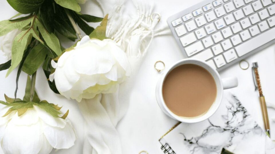 Bild mit Kaffeetasse, weißen Blumen, weißer Tastatur, Kugelschreiber und Notizbuch in hellen Farben