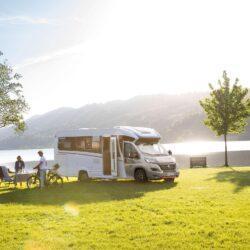 Modern caravan on a meadow with a lake in the background, standing in front of it two people with a bicycle and a folding table