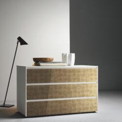 White chest of drawers with golden elements on the stores in front of a white wall next to a floor lamp