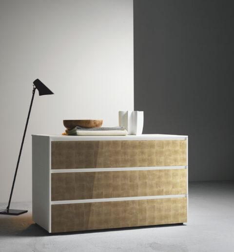 Weiße Kommode mit bronzenen hochglänzenden Ladenfronten vor einer weißen Wand neben einer Stehlampe