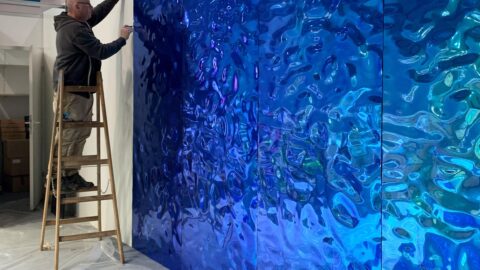 Jemand montiert, auf einer Leiter stehend, auf eine weiße Rückwand blau-grün spiegelnde SIBU DESIGN Dekorpaneele.