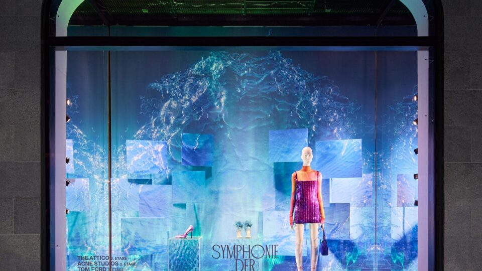 Schaufenster, Hintergund blau mit Wellendruck, die Schaufensterpuppe mit weißem Kopf trägt ein rosa Minikleid. Hauptfarbe des Schaufensters ist blau.