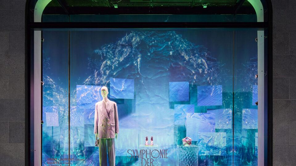 Schaufenster, Hintergund blau mit Wellendruck, die Schaufensterpuppe mit weißem Kopf trägt einen rosaroten Zweireiherblazer. Hauptfarbe des Schaufensters ist blau