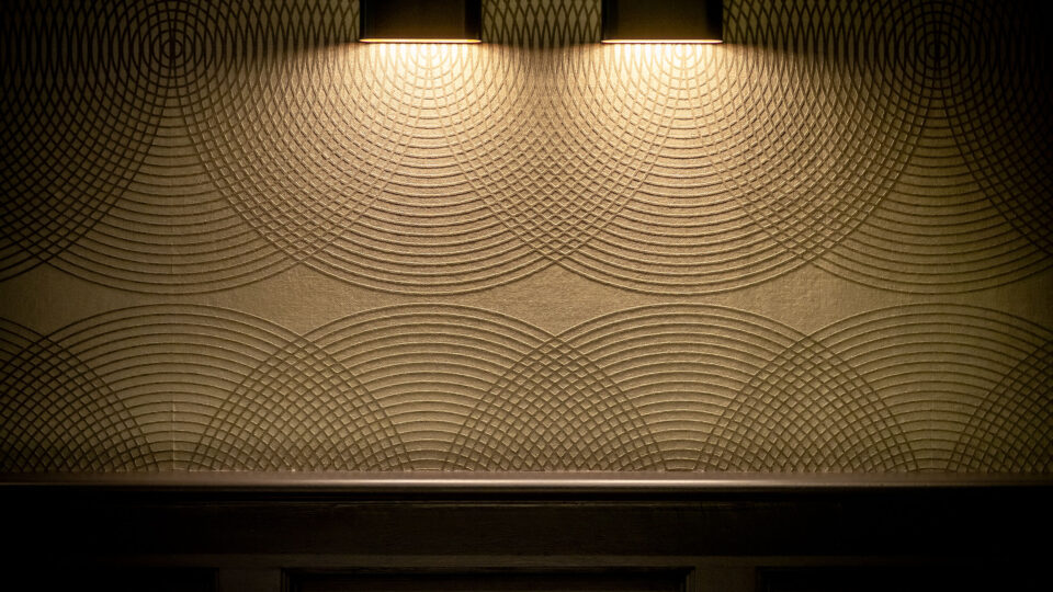 Nahaufnahme einer hölzernen Wandverkleidung, darüber eine Tapete in Beigetönen die Kreislinienzeigt die elegant ineinander laufen. Die Tapete wird durch zwei moderne Spots beleuchtet und die Struktur dadurch perfekt in Szene gesetzt.
