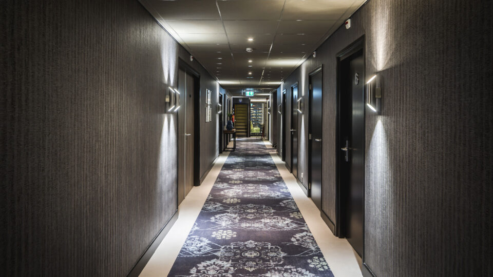 Ein moderner breiter Hotelflur, der verkleidet ist mit einer Tapete die vertikale Linien in verschiedenen Grautönen darstellt