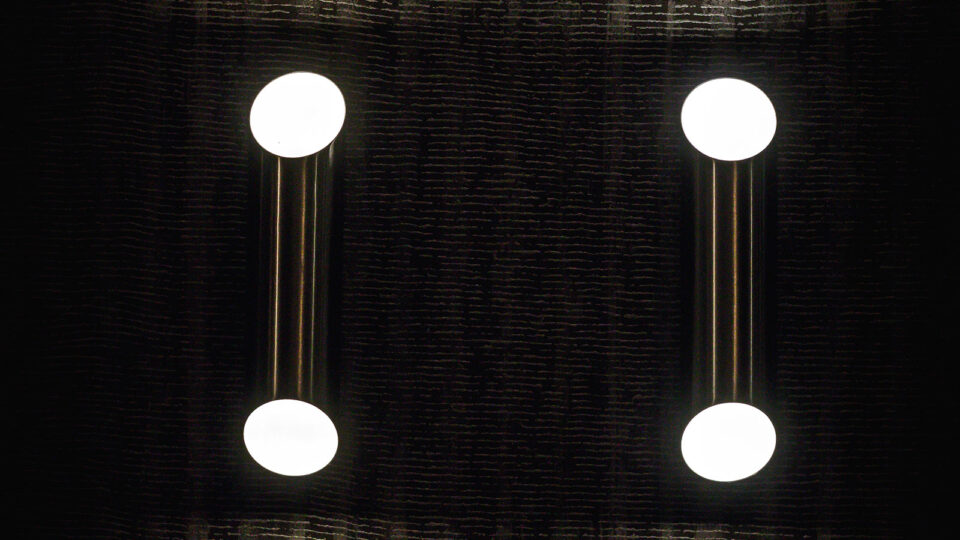 Nahaufnahme von zwei Spots die sowohl nach oben als auch nach unten leuchten und dadurch die dahinterliegende Tapete mit ihren vertikalen Linien und horizontalen Rillen gut in Szene setzt.