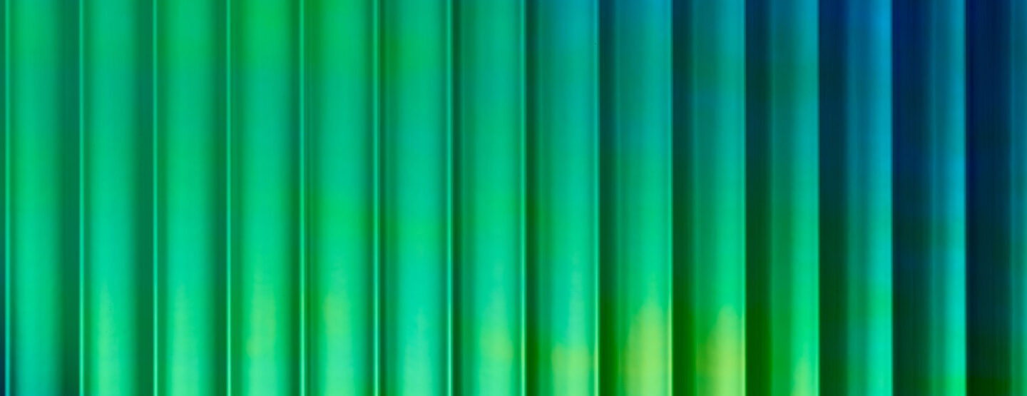 A close-up of a design panel, characterized by wide vertically running lines. The surface is reflective, with a color that shifts from green to blue depending on the viewing angle. The varying heights and depths of the structure create a particularly deep effect that captivates.