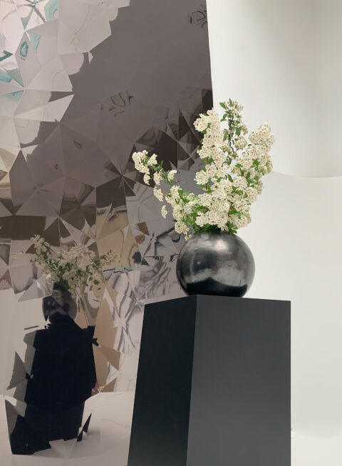 White flowers in a black round vase on a slanted black stool in front of a design panel with a diamond-like structure, anthracite colored and a shiny, reflective surface.