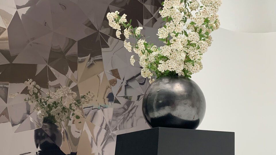 White flowers in a black round vase on a slanted black stool in front of a design panel with a diamond-like structure, anthracite colored and a shiny, reflective surface.