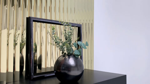 Green leaves and plants in a black round vase, behind which is an empty black picture frame on a black stool placed horizontally. Behind it, a golden SIBU | DESIGN panel.
