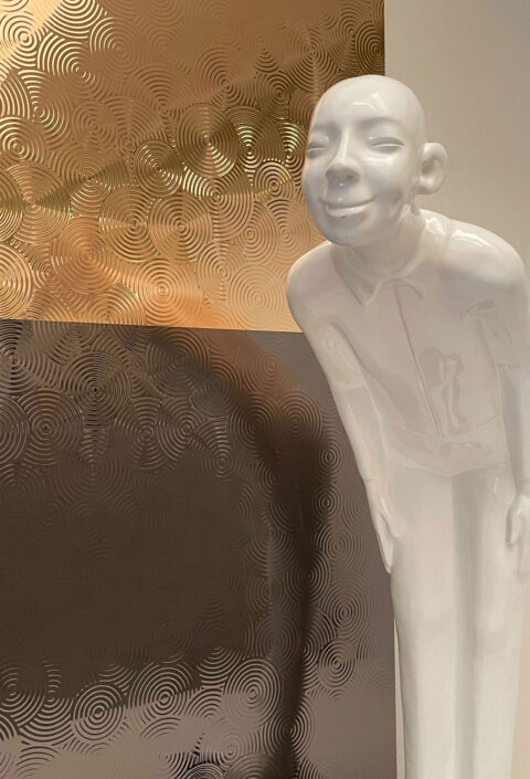 Lächelnde Schaufensterpuppe vor einem strukturierten Designpaneel. Die Struktur zeigt verschieden große Kreise die sich hintereinander zeigen und in sich immer kleinere Kreise beinhalten, Die Oberfläche ist spiegelnd und glänzt im Licht. Die Farbe ist zur oberen Hälfte Gold und zur unteren Hälfte Anthrazit.