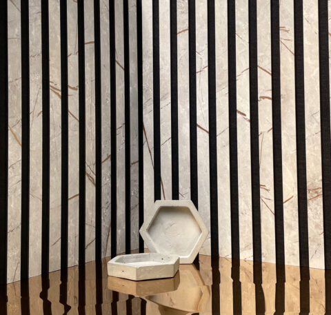 Dargestellt wird ein Akustikpanel, welches in einer Ecke präsentiert wird. Davor ein in Bronze spiegelnder Boden auf dem eine Betonschüssel im Hexagonformat steht.