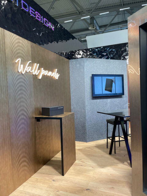 Application photo of a SIBU DESIGN booth at a trade fair. In the background, a scratched, textured, sandy-metallic design panel made of synthetic leather.