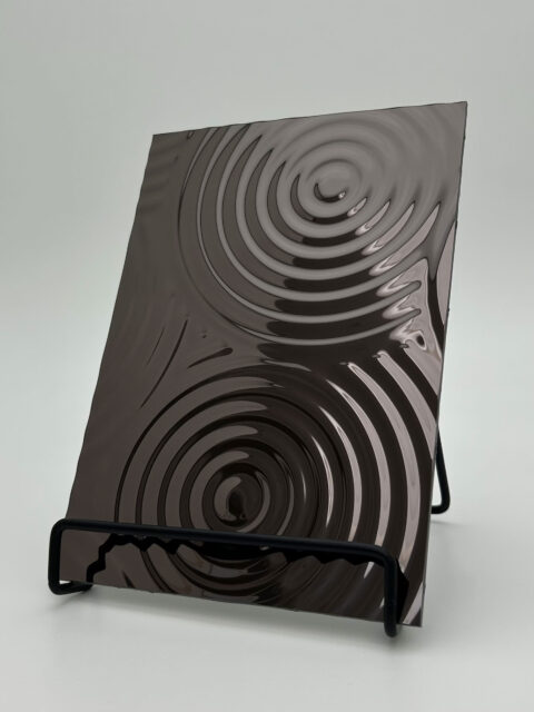 Aufsteller mit einem spiegelnden anthrazitfarbenen Designpaneel, dessen Struktur verschieden große Kreise die sich hintereinander zeigen und in sich immer kleinere Kreise beinhalten, zeigt.